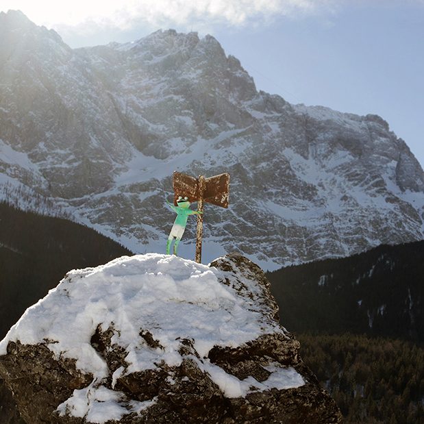 Im Winter friert der Eibsee manchmal zu. Dann werden die kleinen Inseln zu verschneiten Inseln im Eis. Auf einer von ihnen winkt ein Frosch.