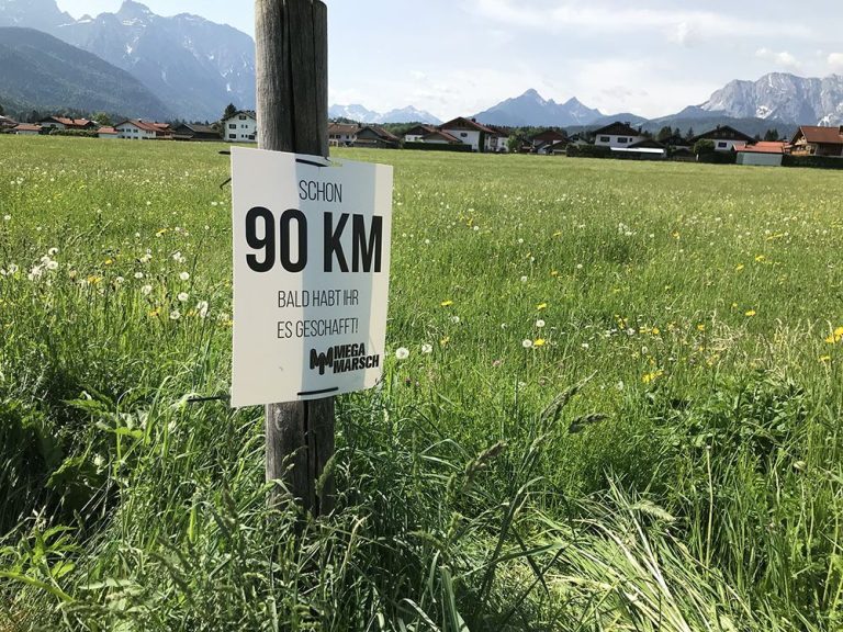 Ein Wegweiser beim Megamarsch München. Er zeigt an, dass schon 90 km gewandert sind – nur noch 10 km bis ins Ziel in Mittenwald.