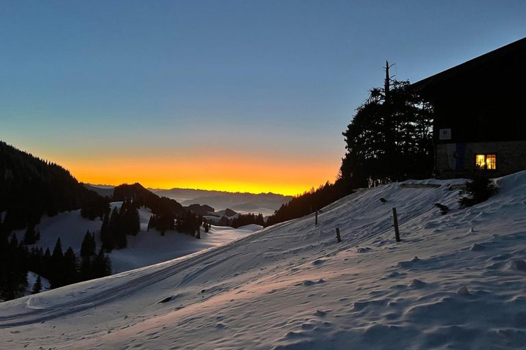 Das Bild zeigt die Priener Hütte im Winter, es liegt viel Schnee. Im Hintergrund ist ein Sonnenauf- oder Sonnenuntergang zu sehen.